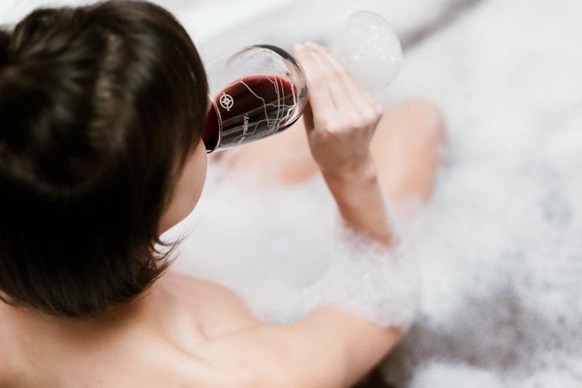 A person drinking from a glass while sitting in a bathtub filled with bubbles. 2