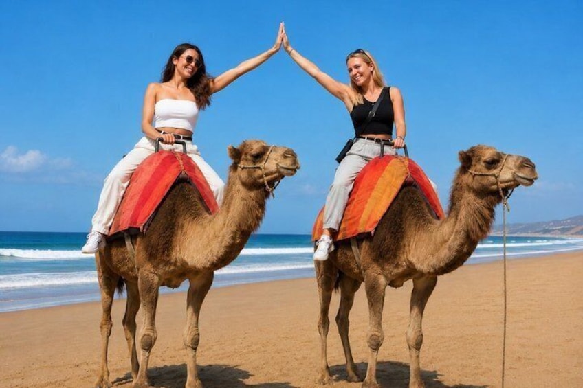 Two women riding camels on a beach with the ocean in the background. 1