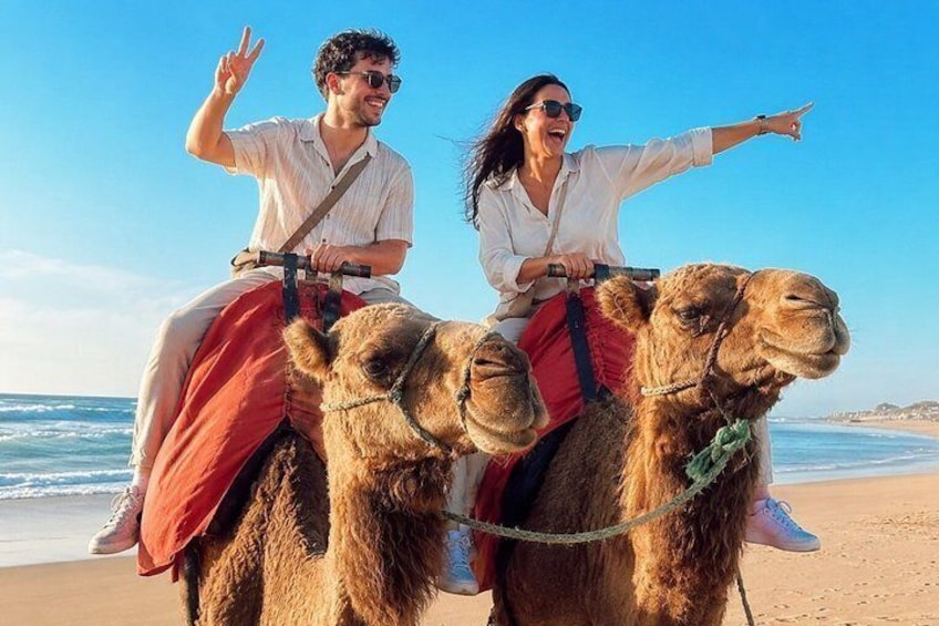 Two people riding camels on a beach with the ocean in the background. 3