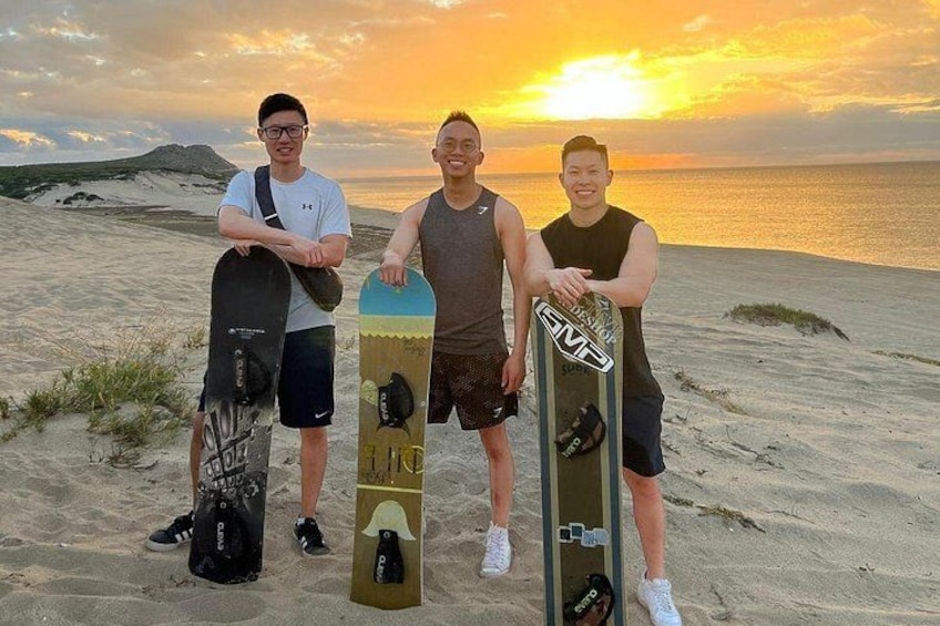 Tre personer står på en strand med snowboard, solnedgang i baggrunden. 3