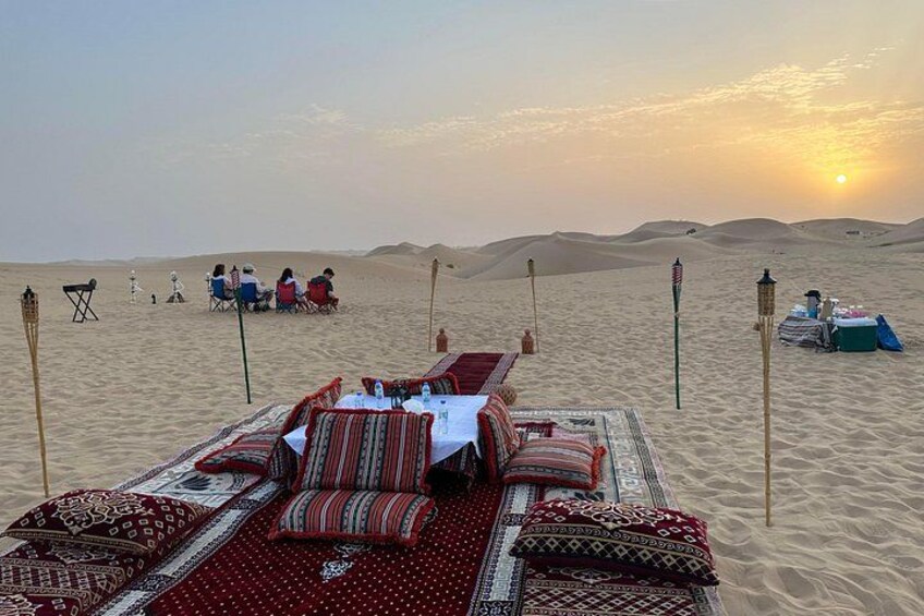 A desert scene with a dining setup featuring a table and chairs, surrounded by sand dunes and a sunset. 1