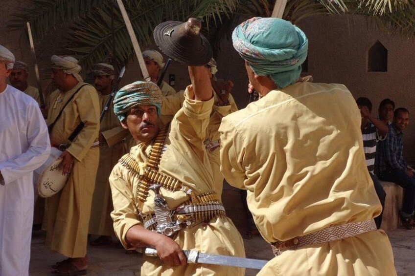 Men in traditional attire, one holding a sword, in a desert setting with palm trees. 1