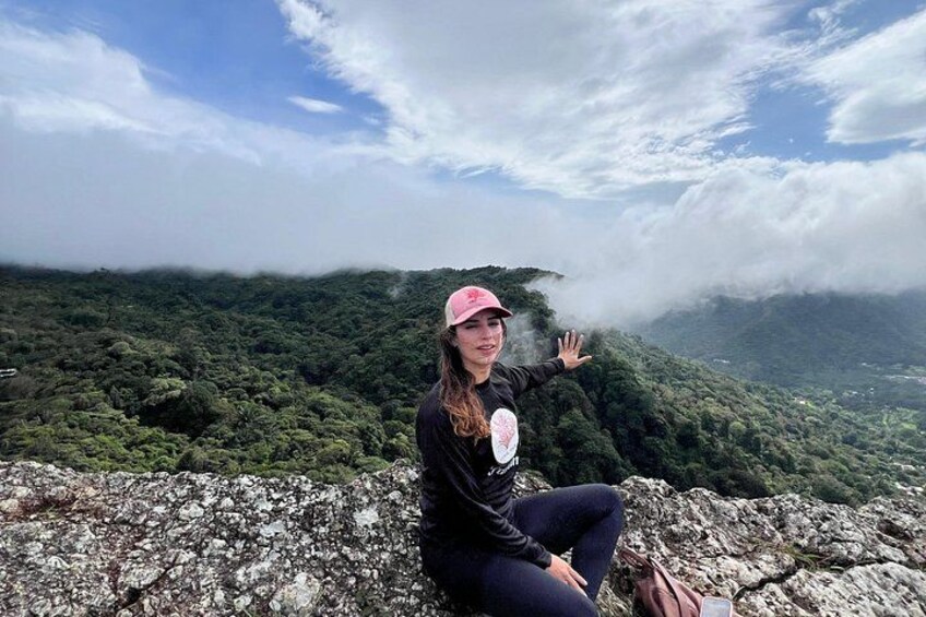 A person sitting on a rocky ledge overlooking a mountainous landscape with dense greenery and clouds. 2