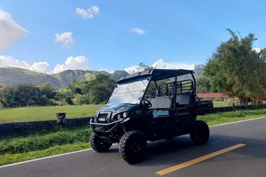 A black off-road vehicle with a roof on a paved road, surrounded by greenery and mountains in the background. 2