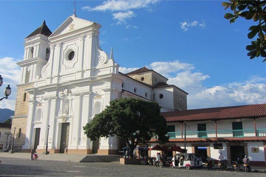 A white church with a bell tower, a tree in front, and a street with people and vehicles. 1