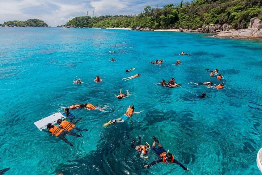 People snorkeling in clear blue water with boats and a rocky shoreline in the background. 1
