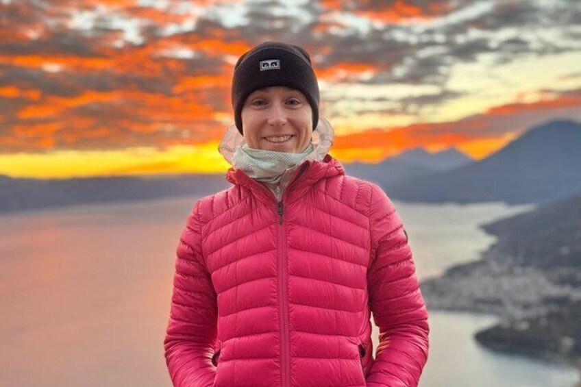 A person in a pink jacket and black beanie standing with hands on hips, overlooking a lake with mountains in the background during sunset. 5