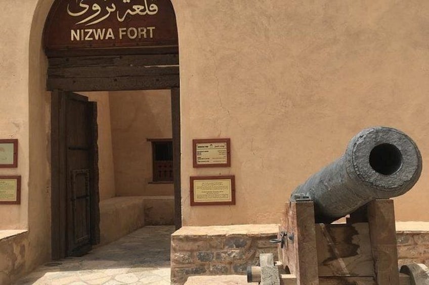 En historisk fortport med en kanon, en skylt med texten ”NIZWA FORT” och informationsskyltar. 3