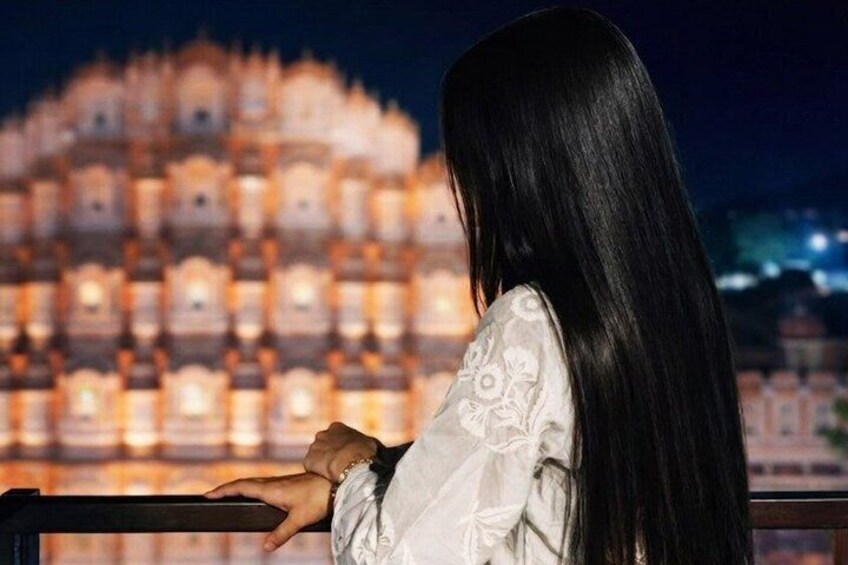 A person with long hair standing by a railing, overlooking a brightly lit, tiered building at night. 1