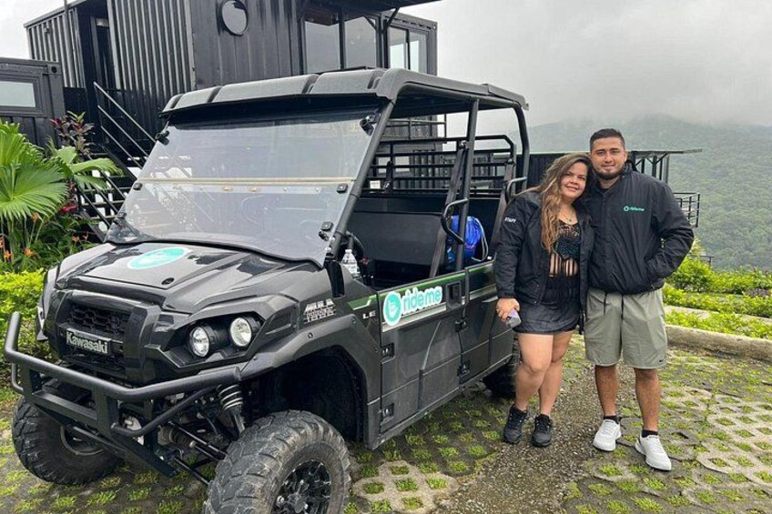 A black off-road vehicle with a logo on the side, parked on a stone path with greenery and a mountainous landscape in the background. 2