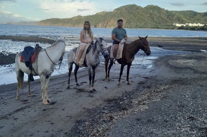 Trois personnes montent à cheval sur une plage, avec une étendue d'eau et des collines en arrière-plan. 4