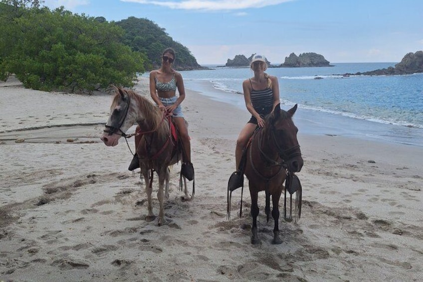 Deux personnes à cheval sur une plage de sable, avec l’eau et la végétation en arrière-plan. 5