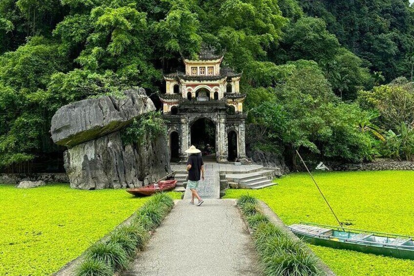 A pathway leading to a traditional gate with a person walking towards it, surrounded by lush greenery and a large rock. 1