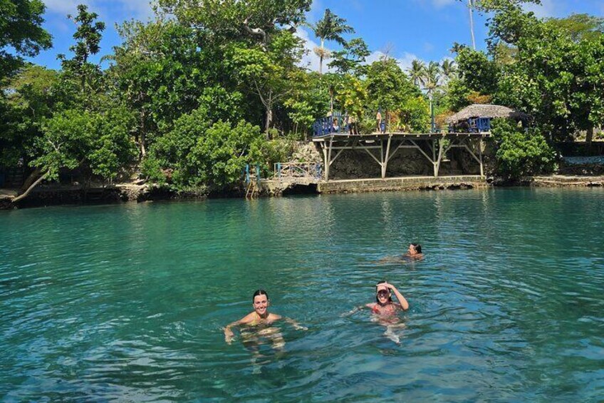 People swimming in a clear blue water body surrounded by lush greenery and a stone bridge. 5