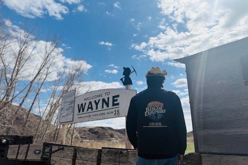 A person standing in front of a 'Welcome to Wayne' sign with a backdrop of a clear sky and mountains. 4