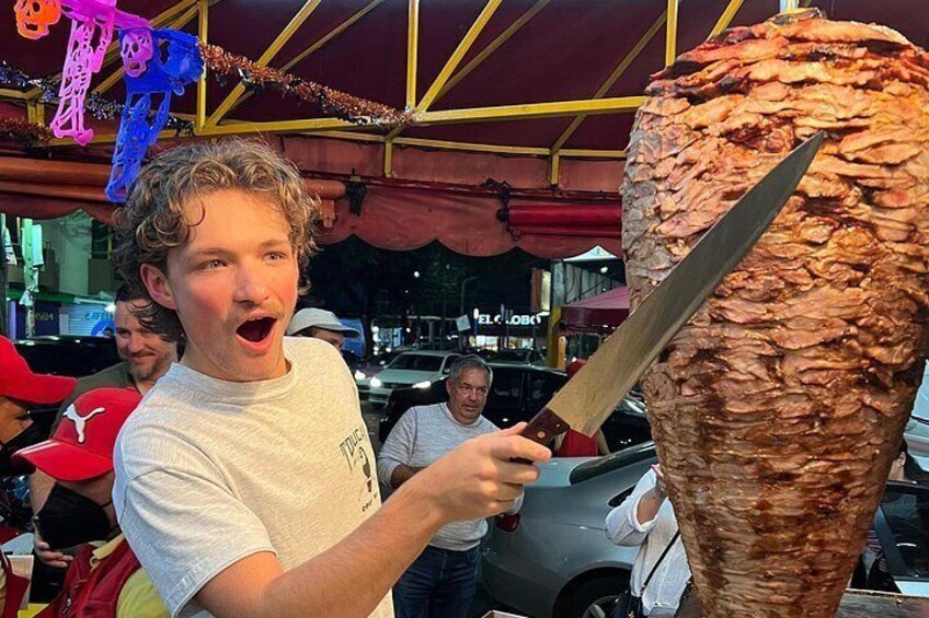 Una persona che tiene un coltello grande vicino a una struttura verticale per la preparazione di cibi. 1