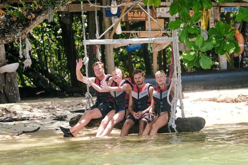 A group of people on a swing in a tropical setting with a wooden structure and greenery in the background. 4