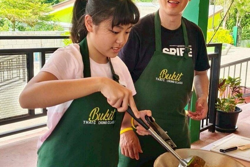 Two people wearing aprons with the text 'Bukit' cooking outdoors. 2