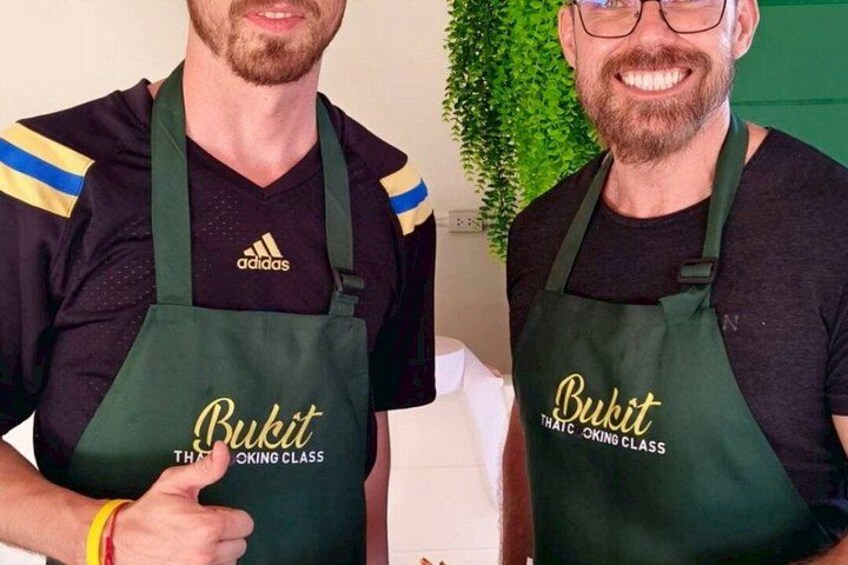 Two individuals wearing green aprons with 'Bukkit' and 'THE MAGIC OKING CLASS' text, standing indoors with a green plant in the background. 5