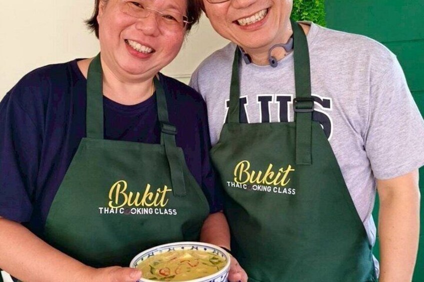 Two people wearing aprons with 'Bukit Thai Cooking Class' text, holding a bowl of food. 5