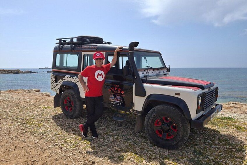 Une personne se tient à côté d’un véhicule tout-terrain blanc et rouge, surmonté d’une planche de surf, garé sur une plage rocheuse avec la mer en arrière-plan. 1