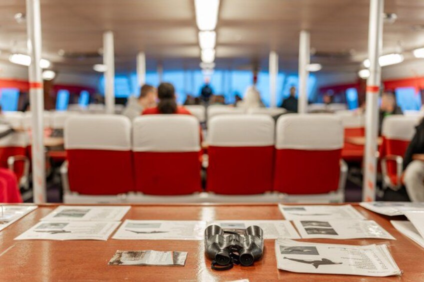 Un coin repas avec des chaises rouges et blanches, une table sur laquelle se trouvent des papiers et un appareil photo, et un arrière-plan flou où l’on aperçoit des personnes. 3