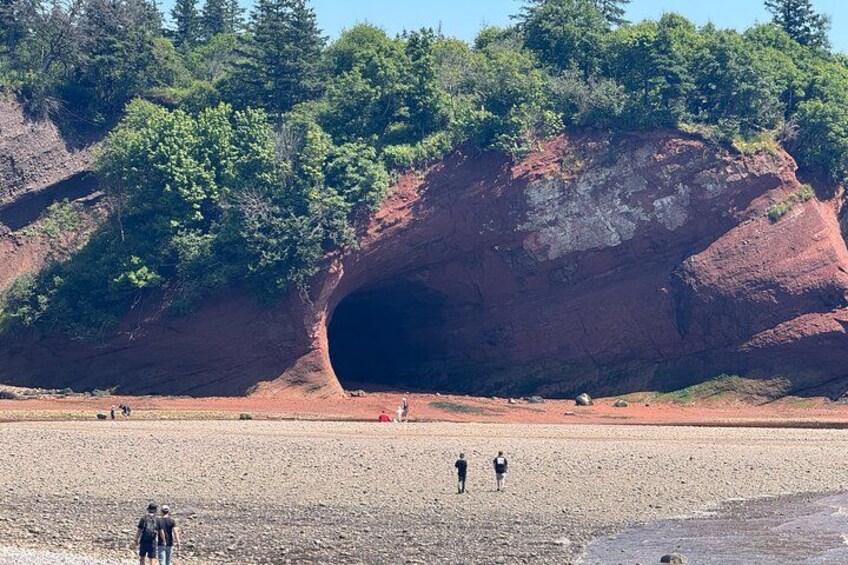 A red rock formation with a cave entrance, a sandy beach, and people walking. 2