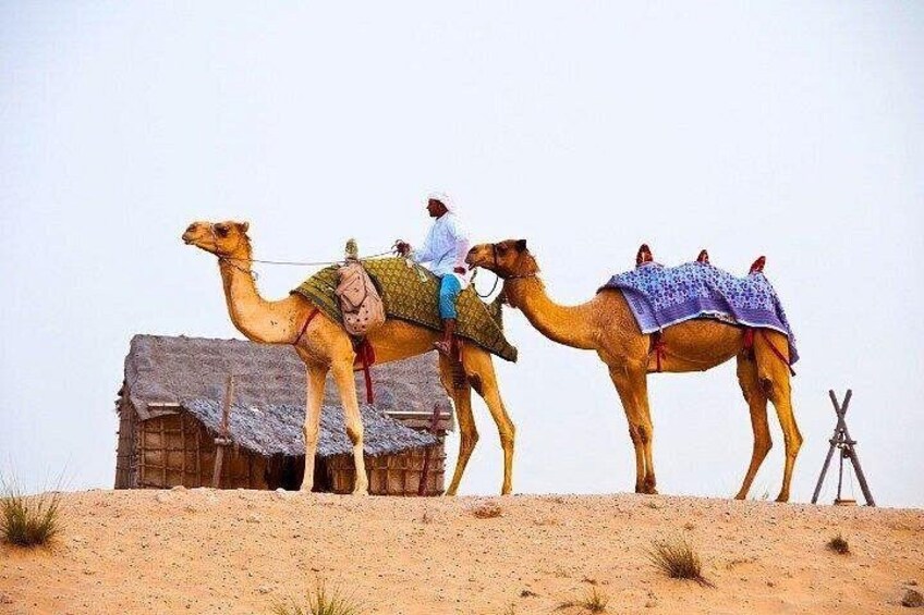 Two camels with riders, a hut, and a tripod in a desert landscape. 5