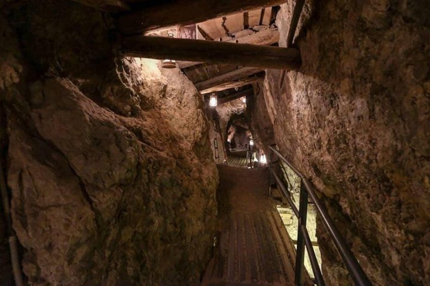 A narrow, dimly lit stone passageway with wooden beams and a metal railing. 4