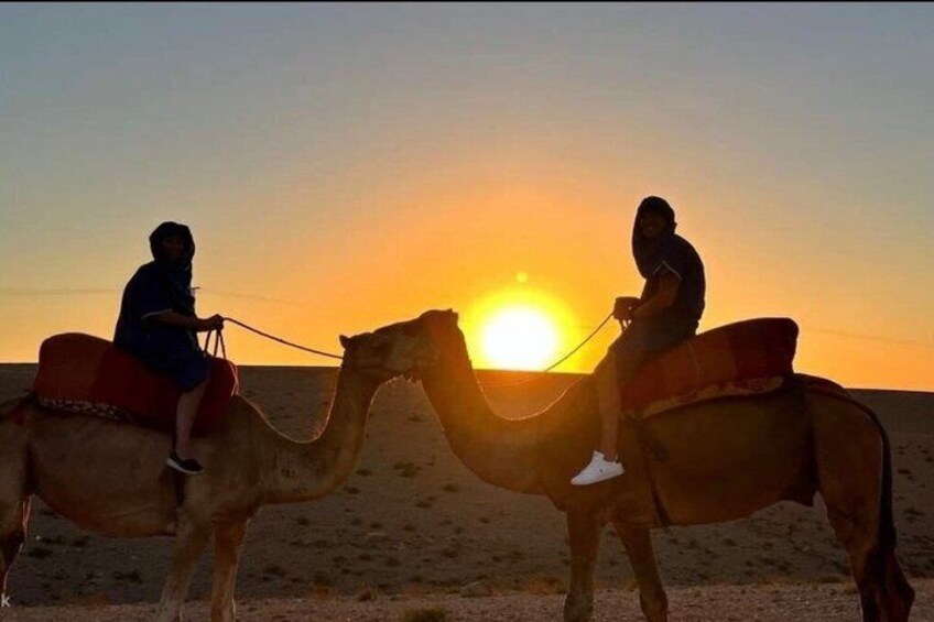 Two people riding camels on a desert path with the sun setting in the background. 2