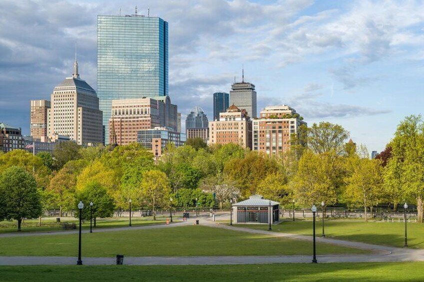 A city skyline with modern and historic buildings, a park with green grass and trees, and a small pavilion. 4