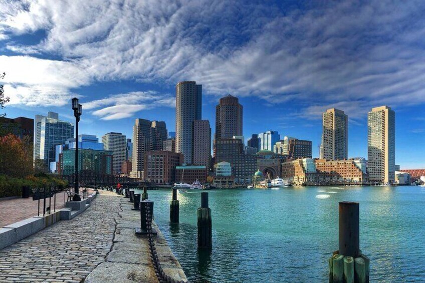 A waterfront cityscape with a cobblestone walkway, tall buildings, and a clear blue sky. 2