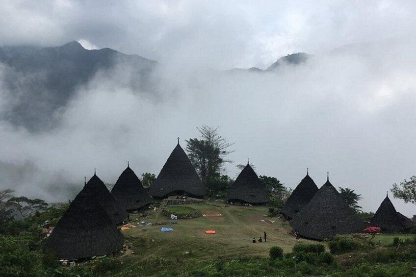 Thatched-roof huts arranged in a semi-circle on a grassy hillside, with misty mountains in the background. 3