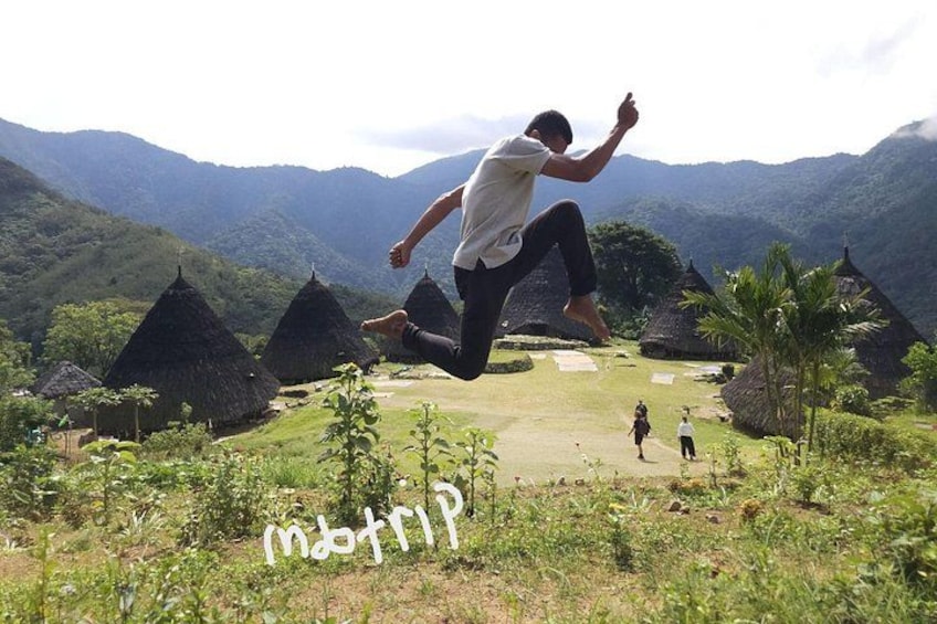 A person jumping in a rural setting with thatched-roof huts and lush greenery. 2