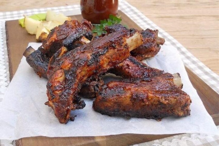 Grilled ribs on a wooden board with a side of sliced avocado and a bottle of sauce. 3