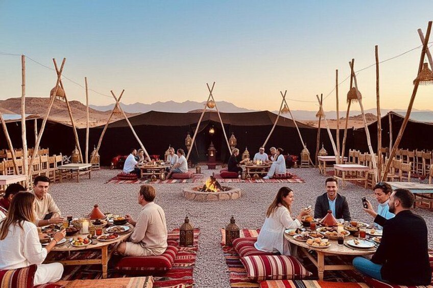 A group of people dining outdoors with tents and a fire in the center. 3