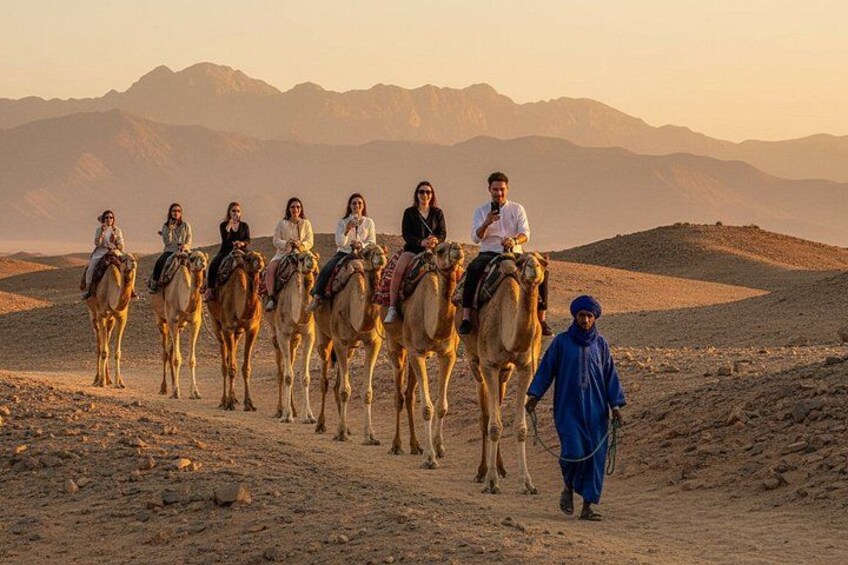 A group of people riding camels on a desert path with mountains in the background. 2