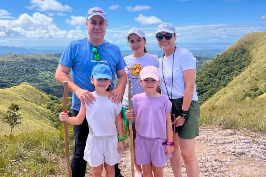 A family of five on a mountain trail with lush greenery and a clear sky. 5
