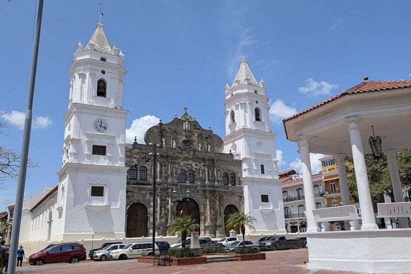 A historic church with two towers, a plaza with cars, and a covered pavilion. 1