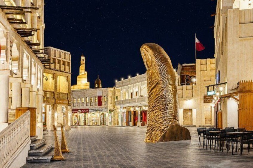 Een verlicht stadsplein 's nachts, met een groot beeldhouwwerk van een vis, gebouwen en een minaret op de achtergrond. 5