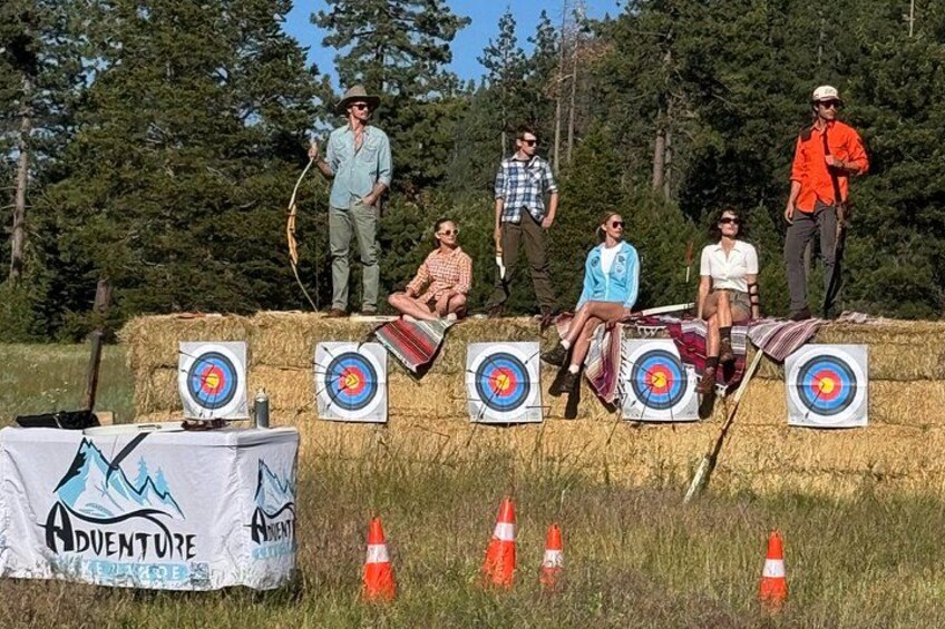 En grupp människor sitter på höbalar med pilar framför sig, troligen vid ett evenemang med bågskytte. 3