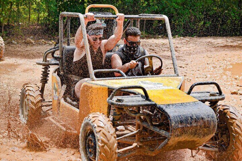 A yellow off-road vehicle driving through a muddy terrain with two people inside. 1