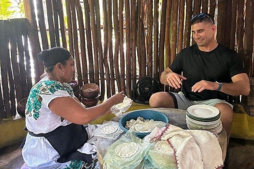 Two people sitting at a table with plates and bowls, a fan, and a wooden fence in the background. 3