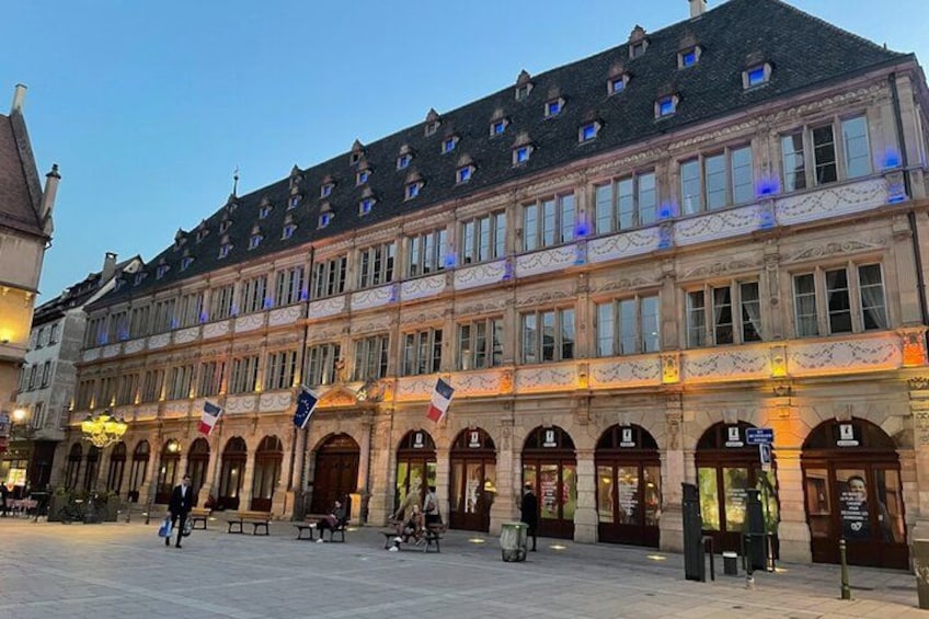 Ein historisches Gebäude mit mehreren Bögen und Flaggen, ein gepflasterter Platz und Bänke. 1