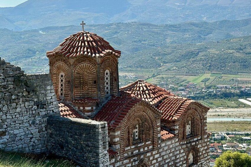 Una iglesia con tejado de tejas rojas y una cruz en la parte superior, rodeada por un muro de piedra y situada frente a un paisaje montañoso. 3