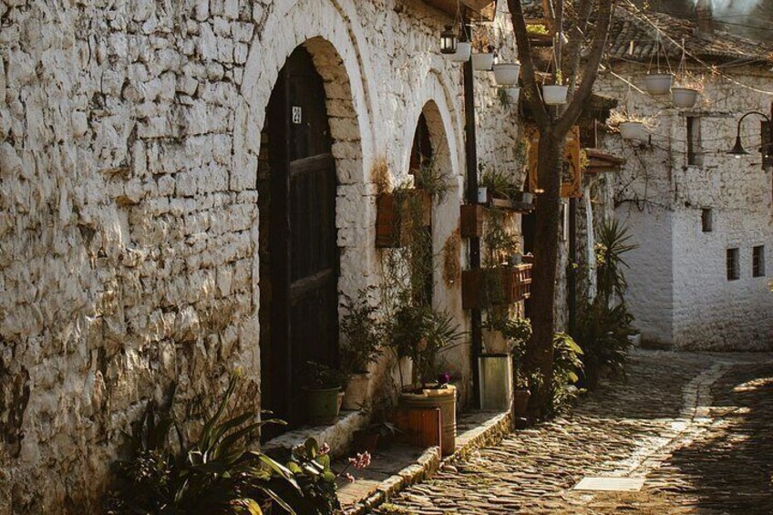 Una strada acciottolata, edifici in pietra bianca, portali ad arco e lanterne sospese, con vasi di piante. 5