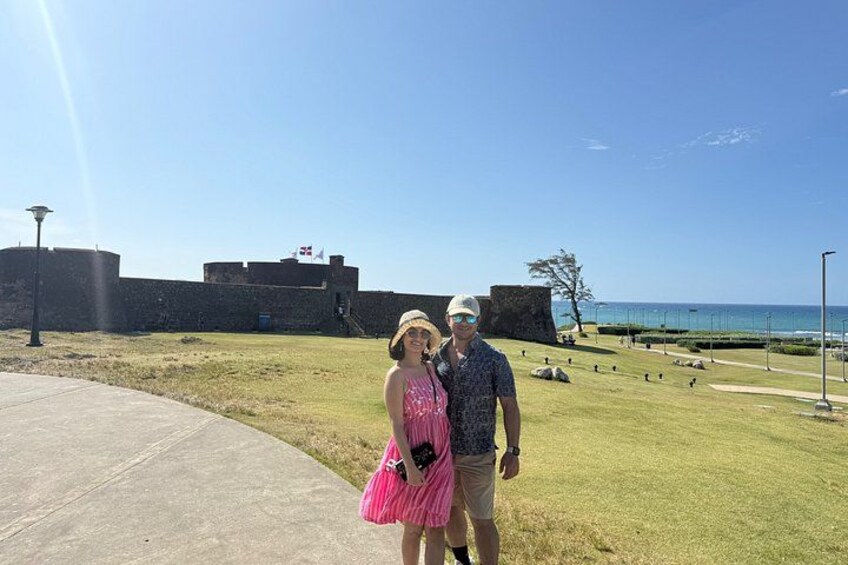 A couple standing in front of a historic fort with a clear view of the ocean and a grassy area with scattered rocks. 3