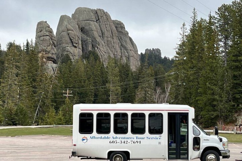 Un autobús turístico blanco con la leyenda 'Affordable Adventures' y un número de teléfono en la carrocería, estacionado en un aparcamiento con montañas al fondo. 1