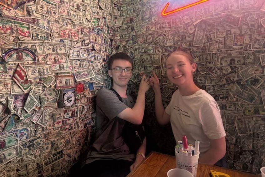 Two people sitting at a table with a wall covered in dollar bills. 5