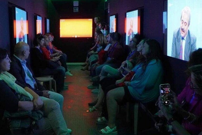 A group of people seated in a dimly lit room, watching a presentation on a large screen. 2